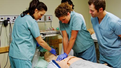 a group of four people gather around a CPR mannequin as one performs CPR 