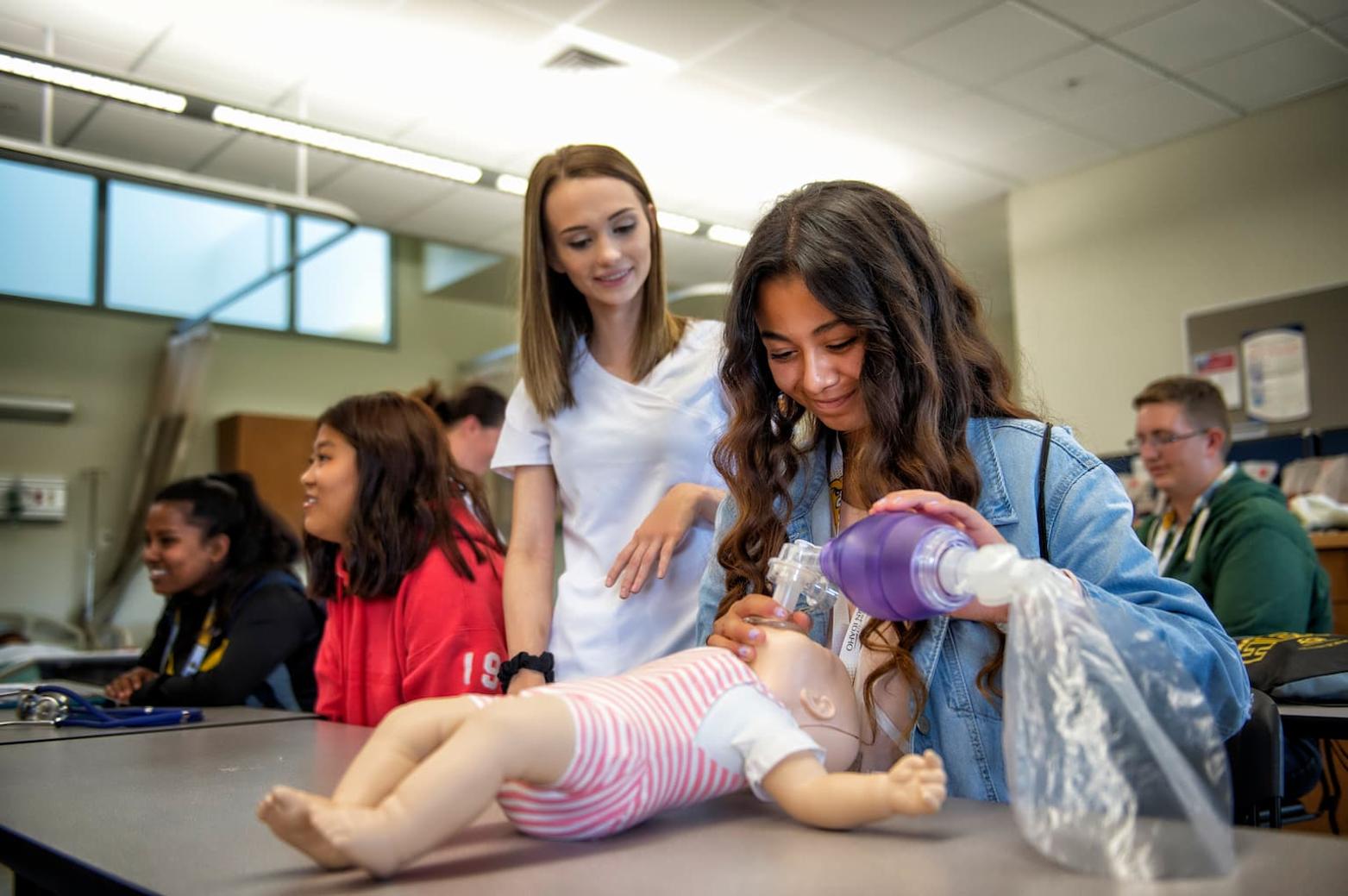 an instructor watches over as a younger person uses a mouth compressor on a CPR mannequin 