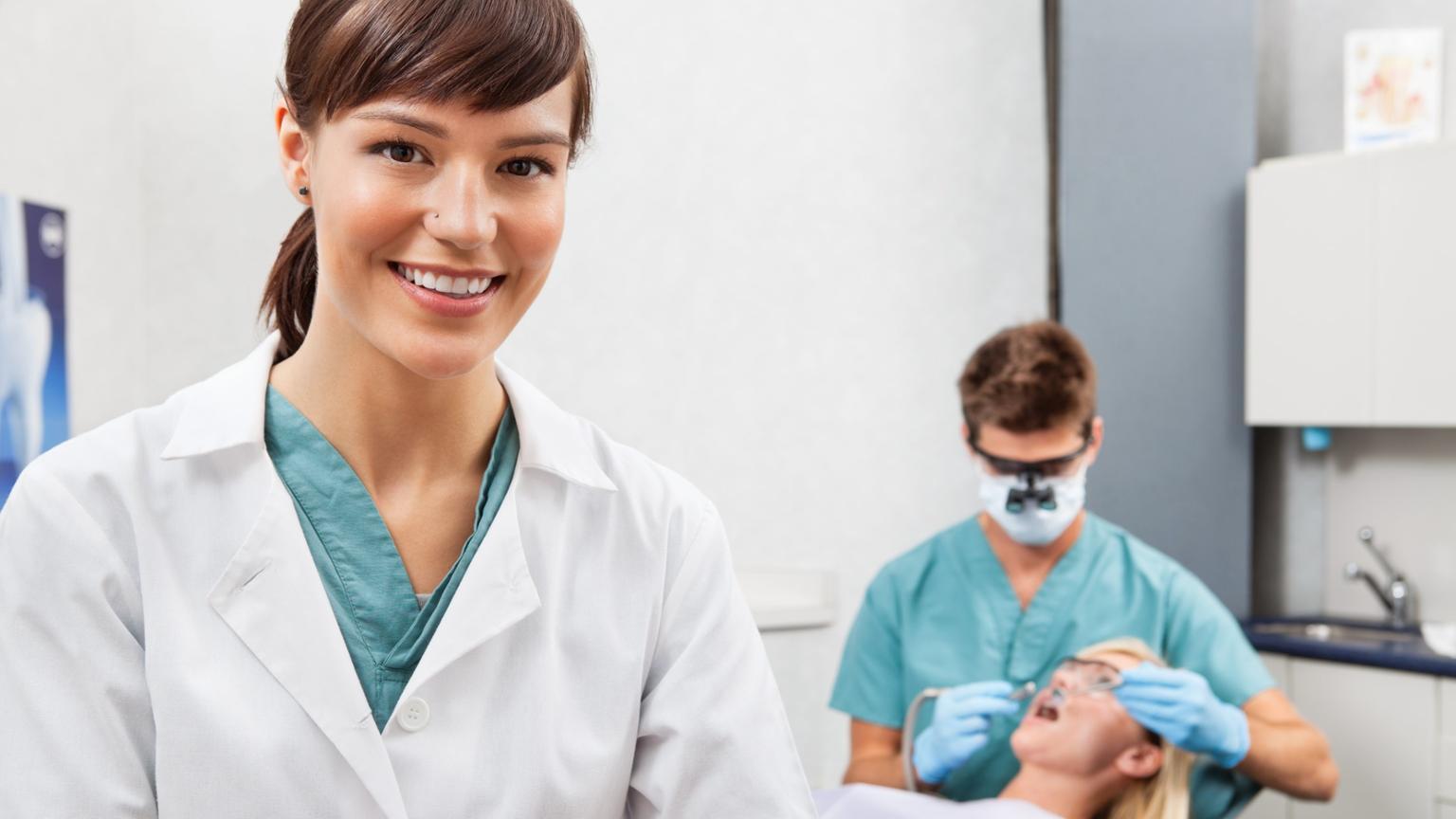 two people in the picture, one is smiling at the camera, the other is in the background wearing a pair of magnifying glasses, working intently on a patient's mouth.