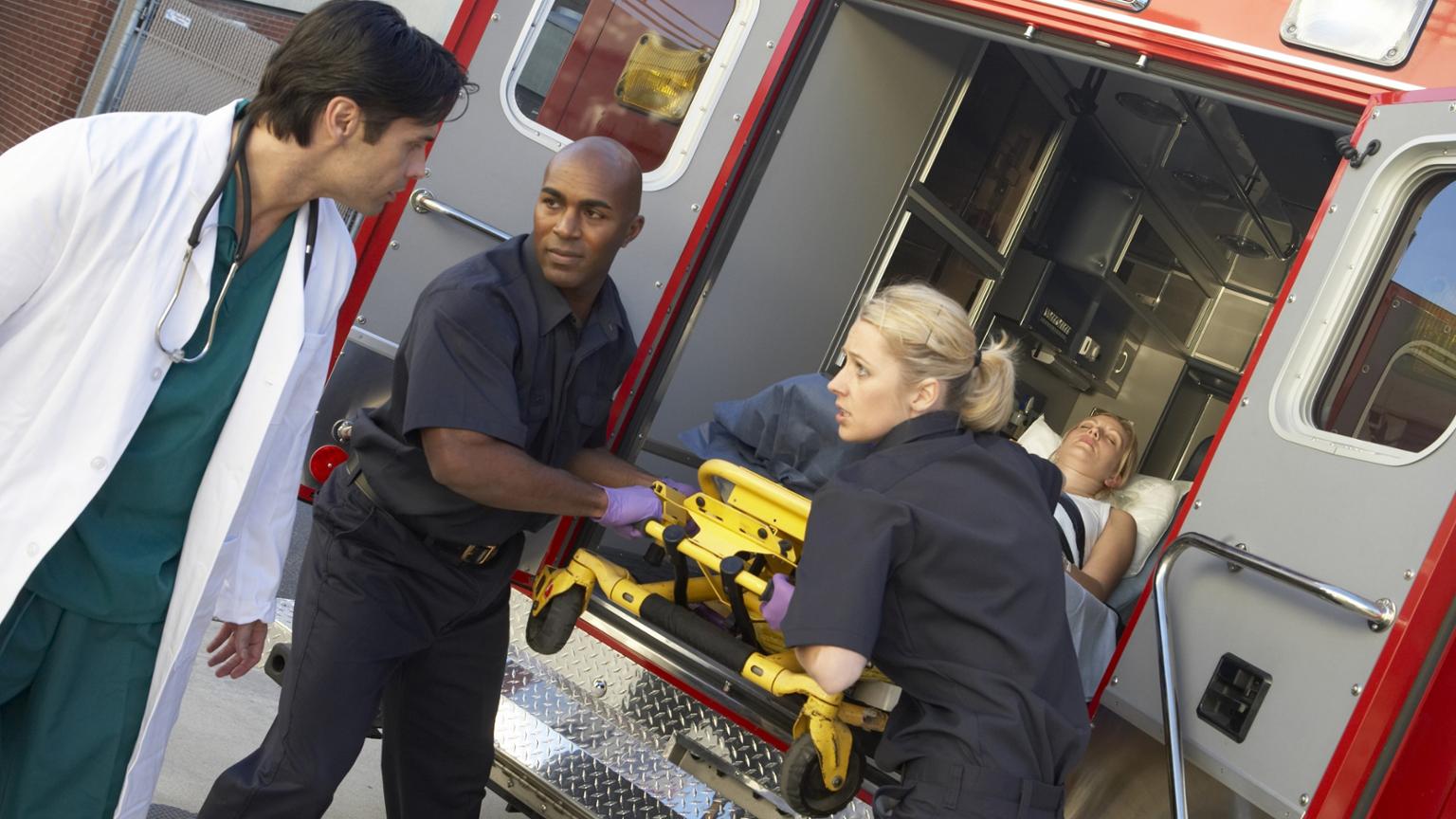four people in the photo, one is in a doctor's coat, two are in EMT uniforms, and one person is laying on a gurney being taken out of an ambulance. 