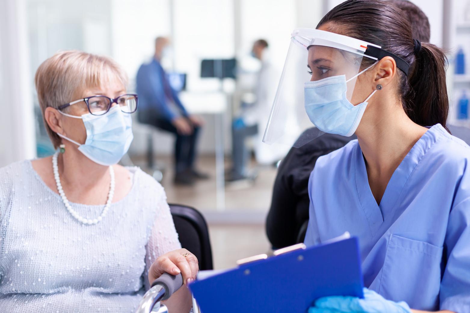 two people sitting next to each other. One is in medical scrubs and they are both wearing masks. 
